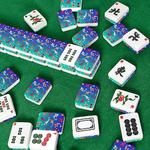 May include: A collection of Mahjong tiles scattered on a green surface. The tiles are white with various symbols and designs, including koi fish patterns in blue and red. Some tiles have Chinese characters.