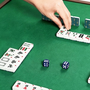 May include: A close-up shot of a Mahjong game in progress. White tiles with red and green markings are arranged on a green felt surface. Two blue dice with white dots are visible. A hand is placing a tile. The game is set on a wooden frame.