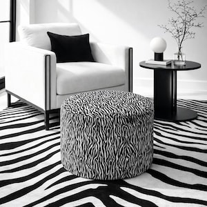 May include: A modern living room scene featuring a white armchair with black trim and a black pillow. A round ottoman with a black and white zebra print sits in front of the chair. A black side table and a zebra-striped rug complete the decor.