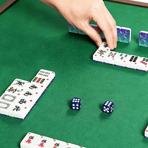 May include: A person's hand arranging white and blue Mahjong tiles on a green felt surface. The tiles have red and green symbols. Two blue dice with white dots are also visible. The game is set on a wooden-framed table.