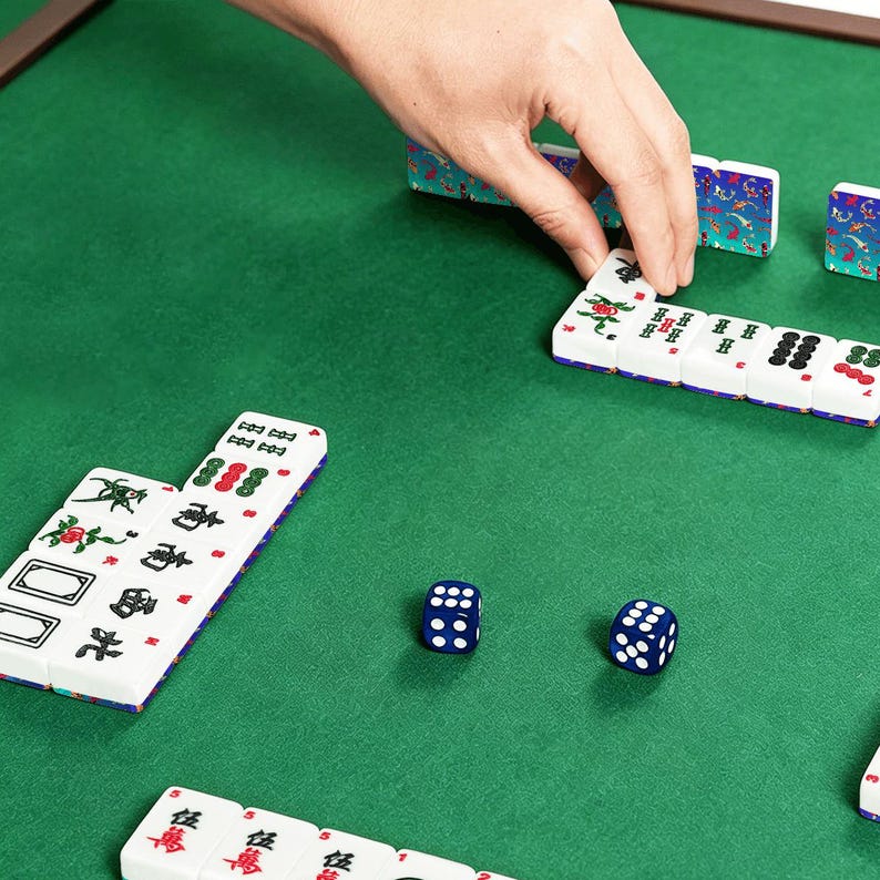 May include: A person's hand arranging white and blue Mahjong tiles on a green felt surface. The tiles have red and green symbols. Two blue dice with white dots are also visible. The game is set on a wooden-framed table.