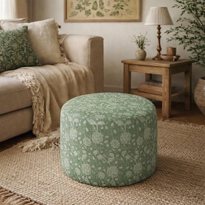 May include: A round, upholstered ottoman with a green floral pattern. The ottoman is the focal point of a living room scene, sitting on a woven jute rug. The room also features a beige sofa, throw pillows, and a wooden side table.