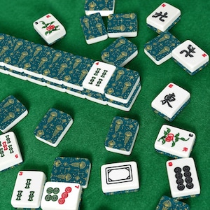 May include: A collection of Mahjong tiles scattered on a green surface. The tiles are white with various symbols and numbers, and some have a teal and gold pattern. The game pieces are arranged in a visually appealing manner.