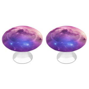 May include: Two round cabinet knobs featuring a vibrant galaxy print with swirling shades of purple, blue, and pink, accented by tiny white star-like specks. Each knob has a smooth, domed top surface and a solid, plain silver-toned metal base.