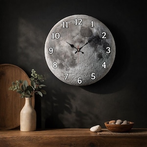 May include: A round wall clock with a detailed moon surface design. The clock face features black hands and white numerals. The clock is mounted on a dark wall, with a wooden shelf and decorative items in the foreground.