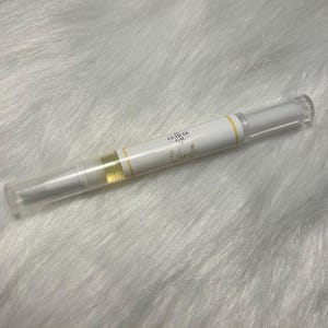 May include: A white cuticle oil pen with a clear cap and a yellow stripe. The pen has the text "The Cuticle Gal Vanilla" printed on it. The pen is lying on a white, fluffy surface.