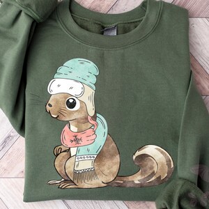May include: An olive green crewneck sweatshirt featuring a graphic illustration of a brown squirrel wearing a light blue winter hat with ear flaps and a matching light blue and pink scarf with a snowflake pattern on the neck area.