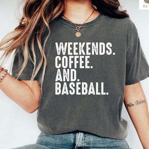 May include: A dark grey t-shirt with the words "Weekends. Coffee. And. Baseball." in white. The shirt is worn with blue jeans and white trainers. The image has a casual, relaxed style.