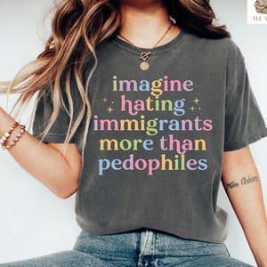 May include: Dark gray t-shirt with the text "imagine hating immigrants more than pedophiles" in colorful letters. The shirt is worn with blue jeans and white sneakers. The image is of a person sitting down.