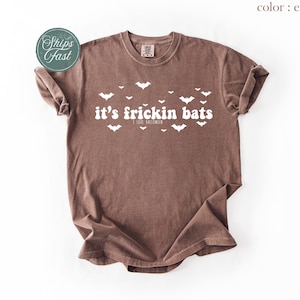 May include: An espresso-colored short-sleeve t-shirt featuring white text that reads it's frickin bats and I love halloween, surrounded by a scattered pattern of small white bat silhouettes across the chest area of the garment.