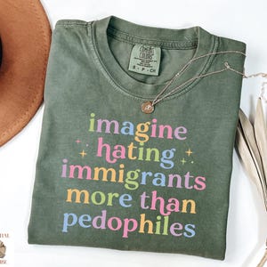 May include: An olive green t-shirt with the text "imagine hating immigrants more than pedophiles" in colorful letters. A gold necklace with a pendant is draped across the shirt. The shirt is folded on a white surface.