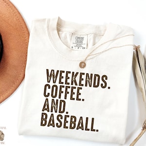 May include: Cream-colored t-shirt with the words "WEEKENDS. COFFEE. AND. BASEBALL." in dark brown block letters. A gold necklace with a pendant rests on the shirt. The shirt is from Comfort Colors and is a size small.