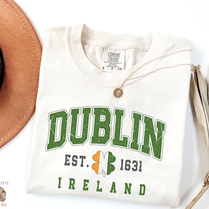 May include: Cream-colored t-shirt with the word "DUBLIN" in distressed green lettering. Below, it reads "EST. 1631 IRELAND" with a shamrock. A gold necklace and a brown hat are visible.