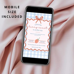 May include: A smartphone displays a baby shower invitation with a strawberry theme. The invitation features the text "someone berry sweet is on the way" on a light pink background with a blue gingham pattern. The phone is on a pink fabric.