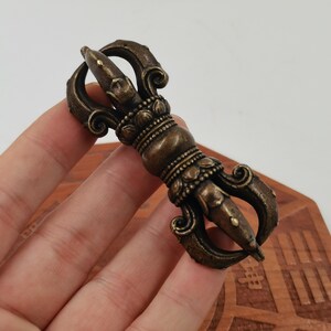 Peut inclure: Un objet rituel vajra en métal de couleur bronze, doté d'une poignée centrale ornée de pétales de lotus et de griffes symétriques aux deux extrémités. La surface présente une patine sombre et usée avec des reflets métalliques visibles.