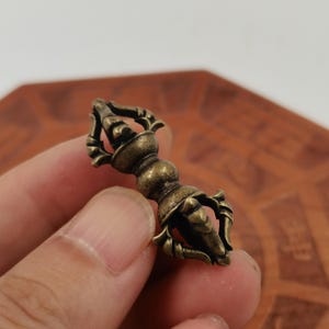 May include: A small, antique-style brass vajra ritual object featuring a central spherical grip flanked by two symmetrical, multi-pronged ends. The metal has a darkened, weathered patina with visible texture and intricate, curved detailing.