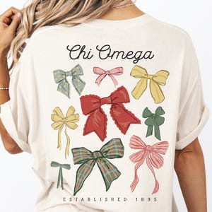 May include: A cream-coloured t-shirt featuring the text Chi Omega at the top and ESTABLISHED 1895 at the bottom. The back of the shirt displays a collection of various illustrated bows in red, yellow, pink, green, and plaid patterns.