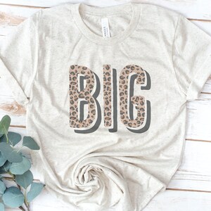 May include: A beige t-shirt with the word "BIG" printed on it in black outlined letters. The letters are filled with a brown and black leopard print pattern.