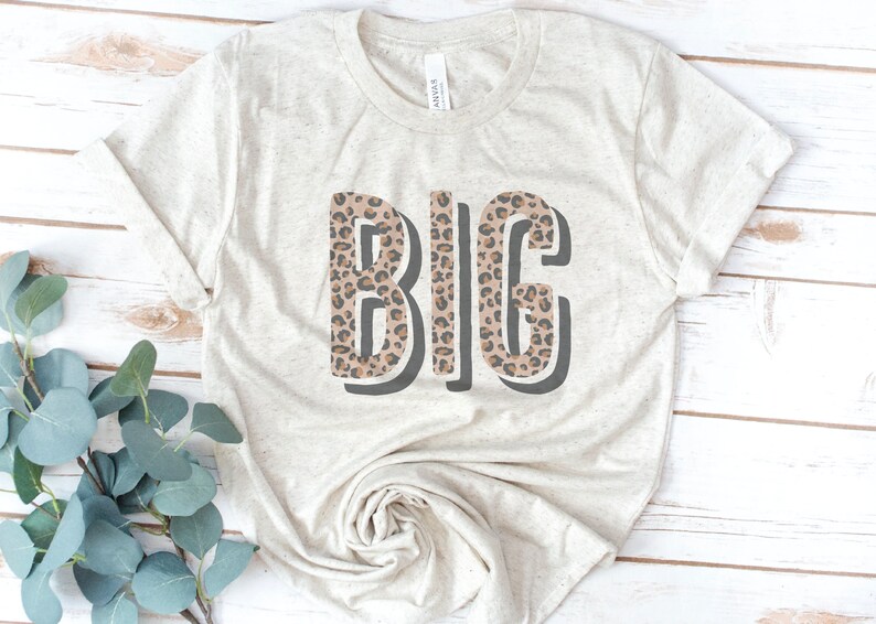 May include: A beige t-shirt with the word "BIG" printed on it in black outlined letters. The letters are filled with a brown and black leopard print pattern.
