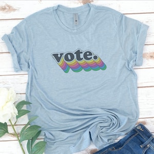 Op de afbeelding: Een lichtblauw t-shirt met een regenboogkleurige afbeelding met de tekst "vote."