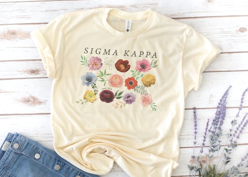 Op de afbeelding: Een cr&egrave;mekleurig T-shirt met een bloemenprint en de tekst "Sigma Kappa" gedrukt op de voorkant.