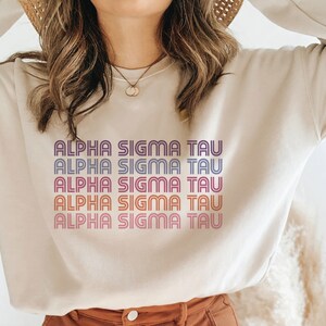 May include: A cream coloured sweatshirt with the text "ALPHA SIGMA TAU" repeated five times in a retro style font. The text is in a gradient of purple, pink, orange, and yellow.