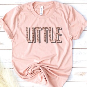 May include: A light pink t-shirt with the word "LITTLE" printed in black outlined letters with a leopard print pattern inside the letters.