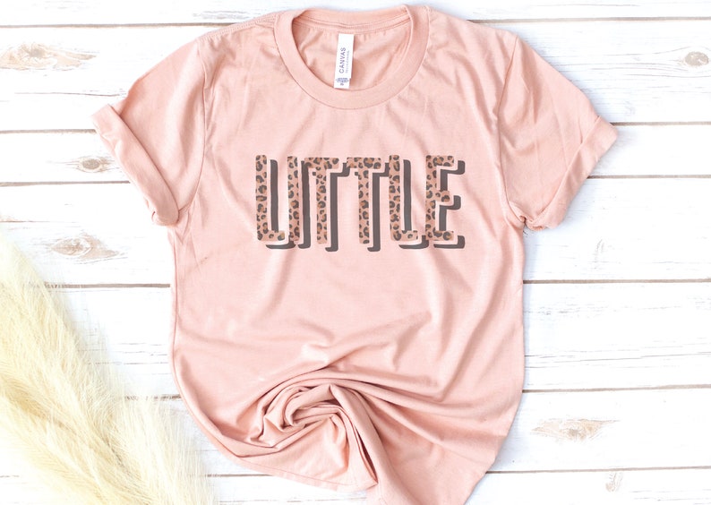 May include: A light pink t-shirt with the word "LITTLE" printed in black outlined letters with a leopard print pattern inside the letters.