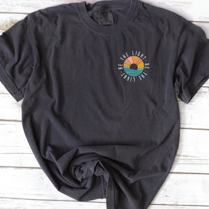 May include: A dark gray t-shirt with a white circle graphic featuring a sunset and the text "Be the light - Be the light".