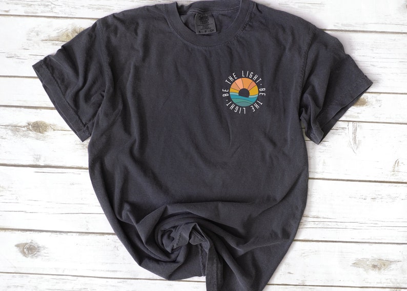 May include: A dark gray t-shirt with a white circle graphic featuring a sunset and the text "Be the light - Be the light".