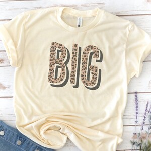 May include: A cream colored t-shirt with the word "BIG" printed in brown and black leopard print.