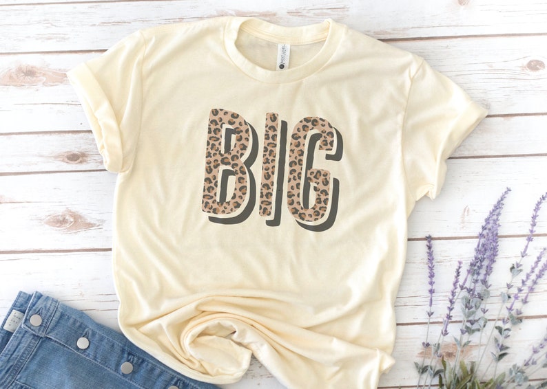 May include: A cream colored t-shirt with the word "BIG" printed in brown and black leopard print.
