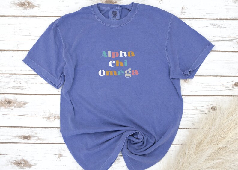Pu&ograve; includere: Una maglietta blu a maniche corte con la scritta "Alpha Chi Omega" stampata in lettere colorate.