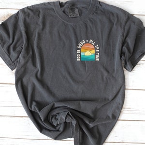 May include: A dark gray t-shirt with a graphic design of a sunset with the text "God is good all the time" in a retro style.