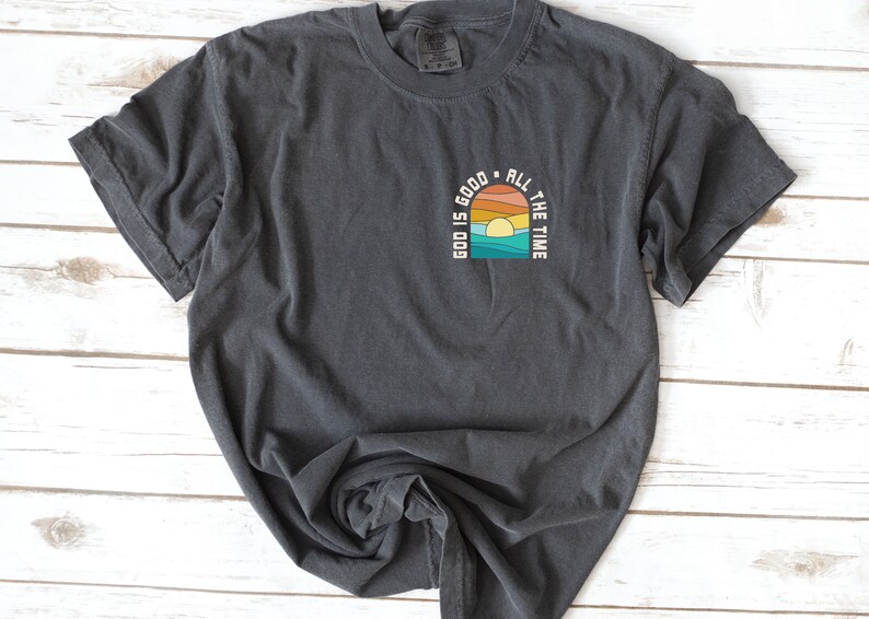 May include: A dark gray t-shirt with a graphic design of a sunset with the text "God is good all the time" in a retro style.