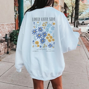 Peut inclure: Sweat-shirt blanc avec un motif floral et le texte "Lambda Kappa Sigma established 1913 October 14 Esse Quam Videri" imprimé au dos.