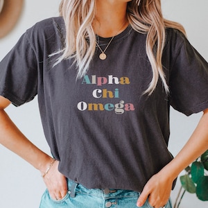 Pu&ograve; includere: Una maglietta nera con il testo "Alpha Chi Omega" in lettere colorate.