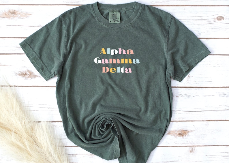 Pu&ograve; includere: Una maglietta verde scuro con le parole "Alpha Gamma Delta" stampate in lettere colorate.
