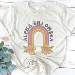 Könnte beinhalten: Ein hellbeiges T-Shirt mit einem Regenbogen- und Blumendesign. Der Text "Alpha Chi Omega" ist in einer Retro-Schriftart geschrieben.