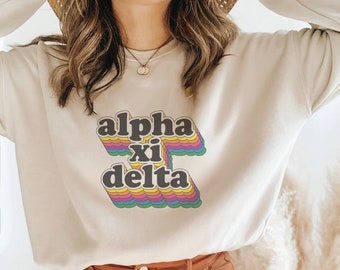 May include: A beige sweatshirt with a rainbow graphic that says "alpha xi delta" in black letters.