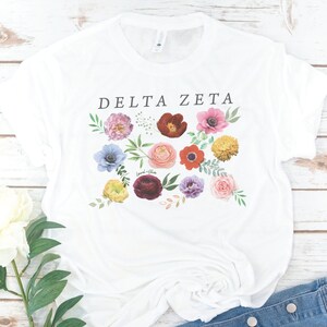 Op de afbeelding: Wit T-shirt met de tekst "DELTA ZETA" in zwart gedrukt. Het shirt heeft een bloemenpatroon met verschillende gekleurde bloemen, waaronder roze, rood, geel, blauw en paars.