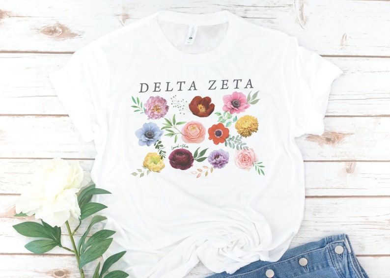 Op de afbeelding: Wit T-shirt met de tekst "DELTA ZETA" in zwart gedrukt. Het shirt heeft een bloemenpatroon met verschillende gekleurde bloemen, waaronder roze, rood, geel, blauw en paars.