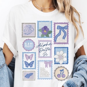 May include: A white short-sleeve t-shirt featuring a graphic print of various postage stamp-style illustrations in shades of blue and purple, including a lion, a flower, butterflies, a seashell, cherries, bows, and the text Alpha Delta Pi.