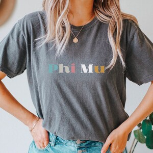 Pu&ograve; includere: Una maglietta grigia scura con il testo "Phi Mu" in lettere colorate. Le lettere sono in un font in stile retr&ograve;.