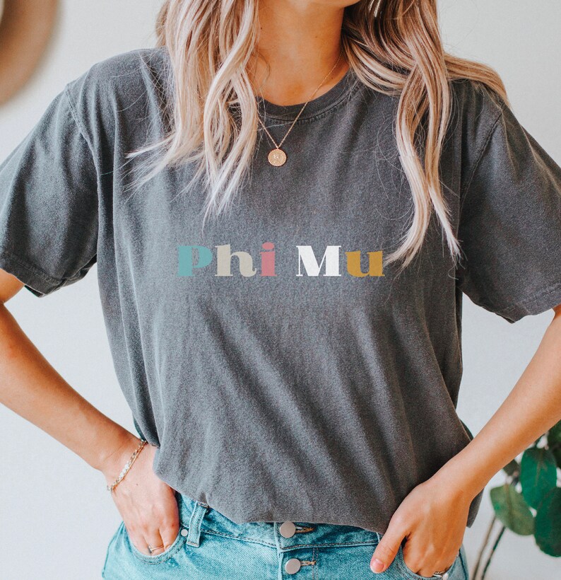 Pu&ograve; includere: Una maglietta grigia scura con il testo "Phi Mu" in lettere colorate. Le lettere sono in un font in stile retr&ograve;.