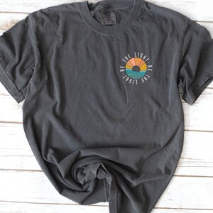 May include: A dark gray t-shirt with a circular graphic design featuring a sunset and the text "Be the light. Be the light."