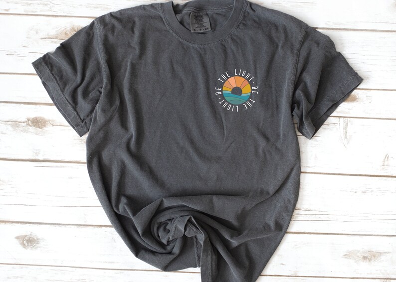May include: A dark gray t-shirt with a circular graphic design featuring a sunset and the text "Be the light. Be the light."