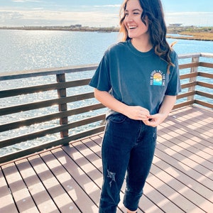 May include: A woman wearing a dark blue t-shirt with the text "God is good all the time" and a sunset graphic. She is wearing dark blue jeans and black and white sneakers. She is standing on a wooden pier with a body of water in the background.