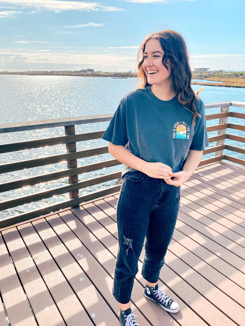 May include: A woman wearing a dark blue t-shirt with the text "God is good all the time" and a sunset graphic. She is wearing dark blue jeans and black and white sneakers. She is standing on a wooden pier with a body of water in the background.