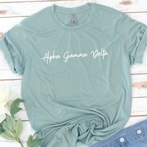 Puede incluir: Una camiseta azul claro con el texto "Alpha Gamma Delta" escrito en cursiva blanca.